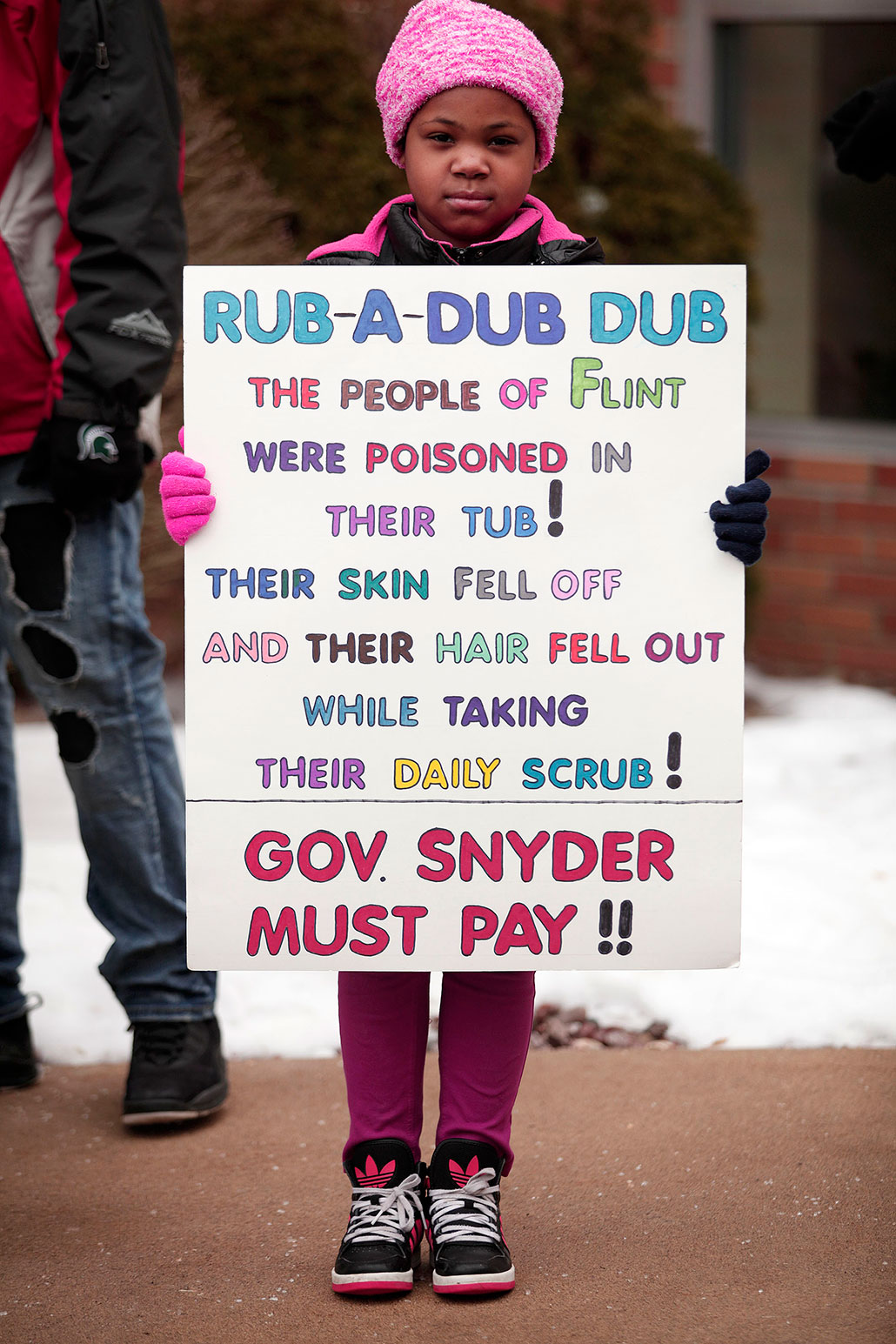 a photo of a young Black girl wearing a pink hat holding a poster that reads 'RUB-A-DUB DUB The people of Flint were poisoned in their tub! Their skin fell off and their hair fell out while taking their daily scrub! Gov. Snyder must pay!'