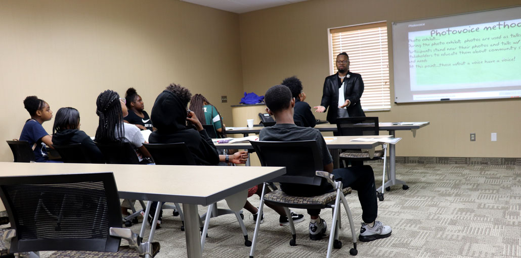 a photo fo Kent Key, a Black man in a blazer, standing in front of a classroom and presenting to a group of Black teenagers