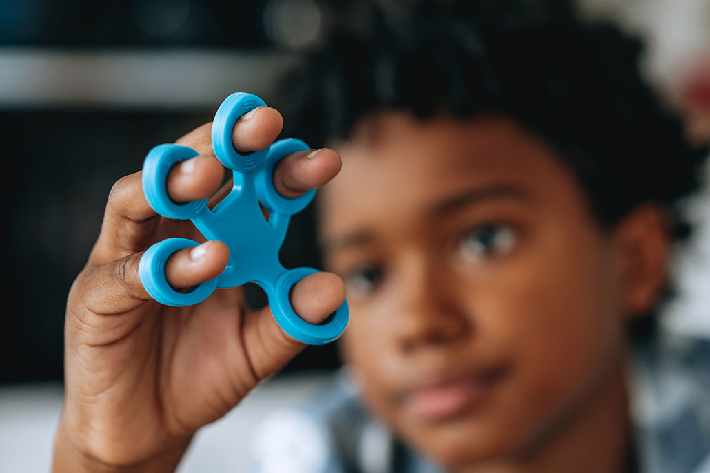 A view of a kid holding and playing with a blue squishy fidget in front of his face. His hand is in focus in the foreground. His face is blurred in the background. He has brown skin and dark curly hair and looks absorbed in playing with his toy.