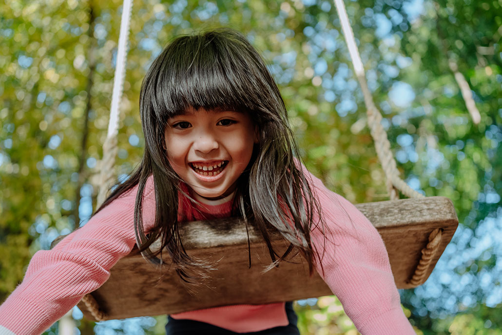 A young girl with pale skin and dark brown hair is smiling and swinging on her stomach on a swing, behind and above her are green leaves and blue sky. 