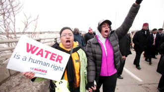 A photo of people marching across a bridge over the Flint River from February 19, 2016. A Black woman in the middle of the image is holding hands with another Black woman, both are raising their voices in protest. The woman on the left has a sign that reads 'Water is a human right' - MI Nurses Association. Behind the women and to the right other protesters can be seen marching.