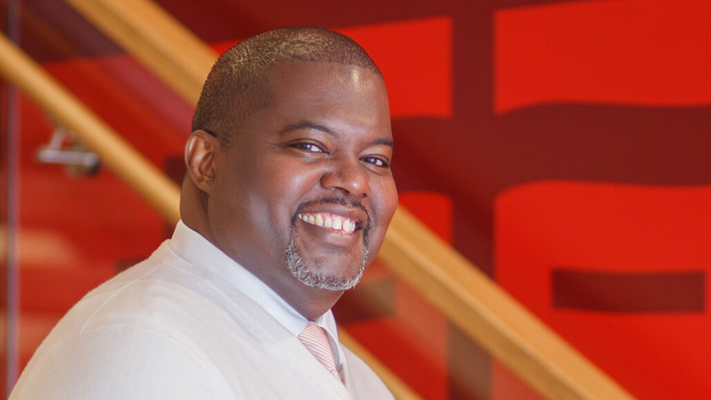 A photo of Kwesi Joseph, a Black man with a big smile, short hair and a neatly trimmed beard. He's wearing a dress shirt with a sweater and a pink tie.