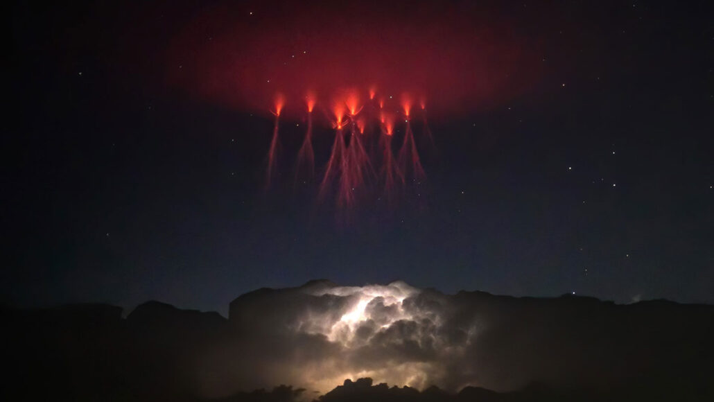 a cluster of what look like red lightning bolts appear in the sky above a storm cloud lit up by lightning