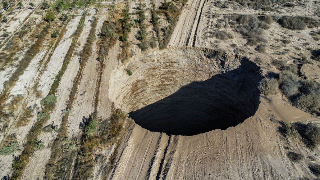 an aerial view of a giant circular sinkhole in a desert with light tan sand. The bottom of the hole is not visible and it seems very deep.