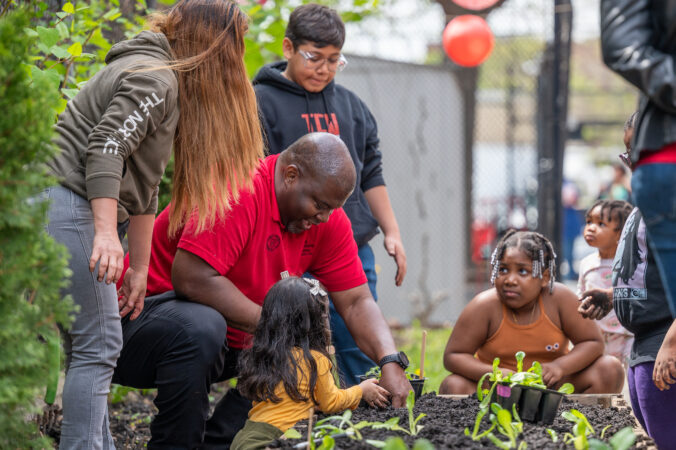 Kwesi Joseph kneels in front of a plot of soil. He is wearing a red polo shirt and dark pants. He is looking down and has one hand in the soil. He is helping a young girl with long black hair with the plot of soil. Two girls and one boy stand next to them. 