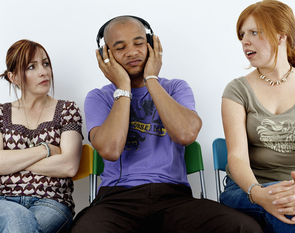 two women stare in dismay at a man sitting between them listening to headphones that are too loud. The man is absorbed in his music and ignoring them