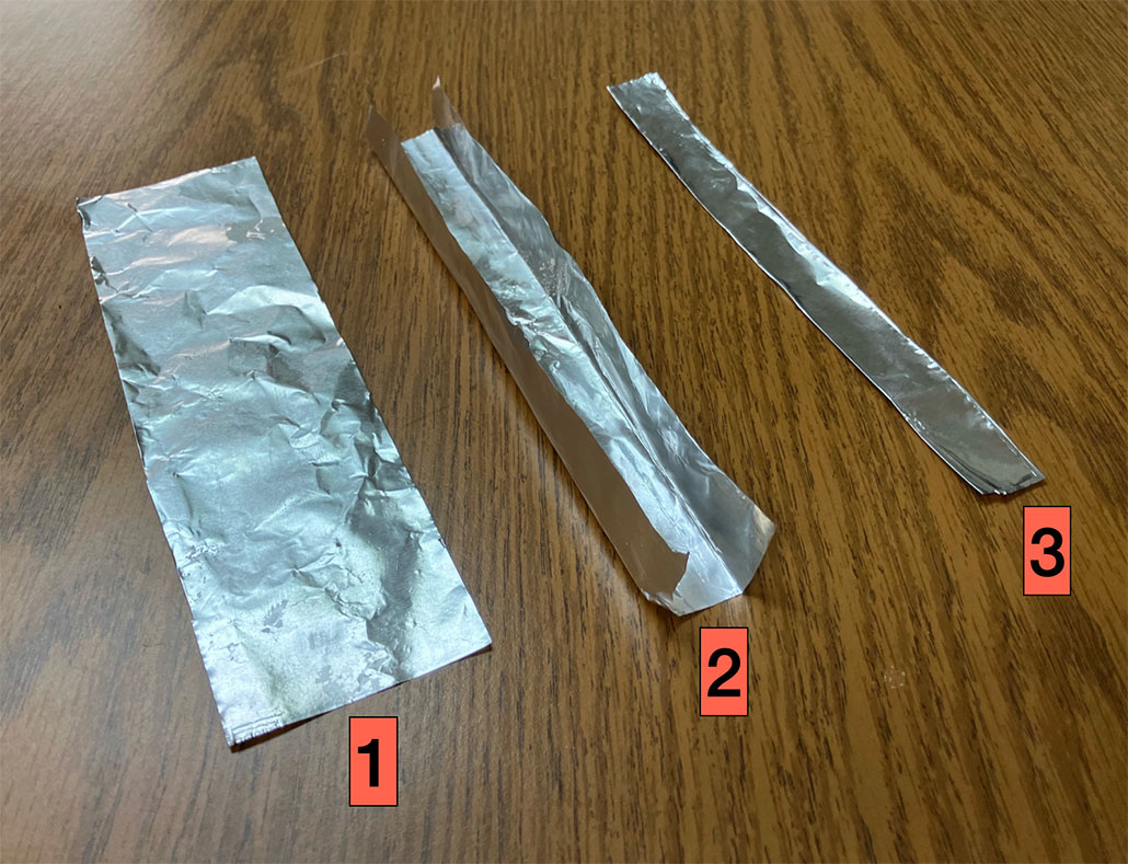 three strips of aluminum foil lay on a desk: one lying flat, a second with its two sides folded lengthwise and bent upward, the third with those two upward-bent flaps laid flat so that the strip is folded three times lengthwise 