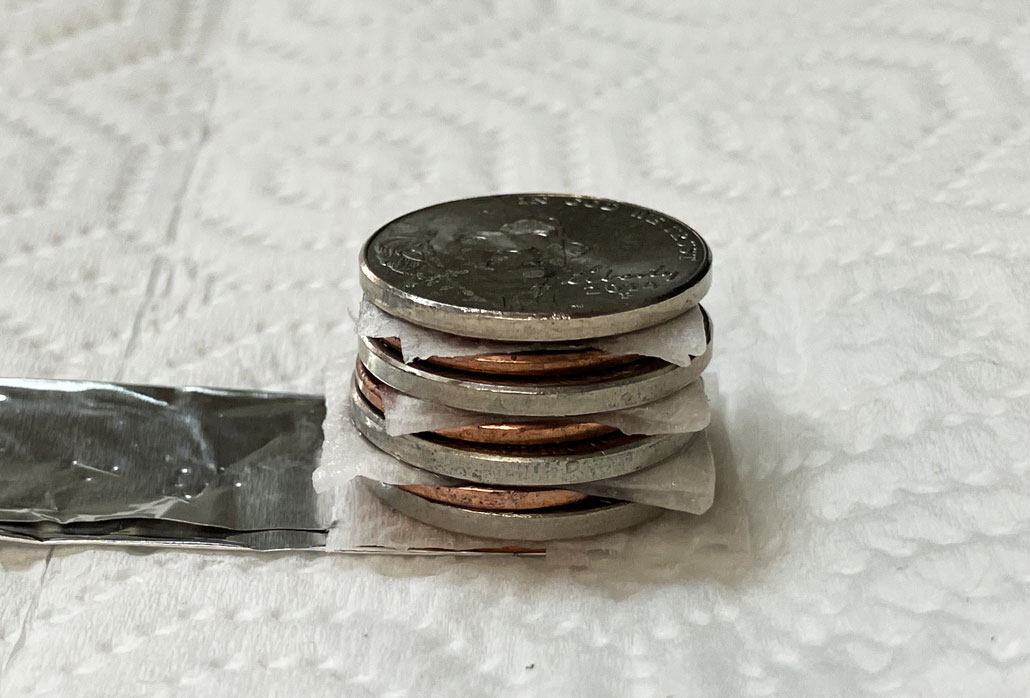a voltaic stack made of alternating pennies, nickels and damp sheets of paper towel sits atop a strip of aluminum foil atop a dry paper towel