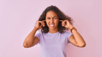 a woman with medium brown skin stands against a purple background. She is wearing a lavender shirt and grimacing while putting her fingers in her ears, as though her surroundings are painfully noisy.