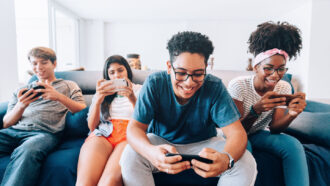four teens sit on a couch all smiling at something they're watching on their phones