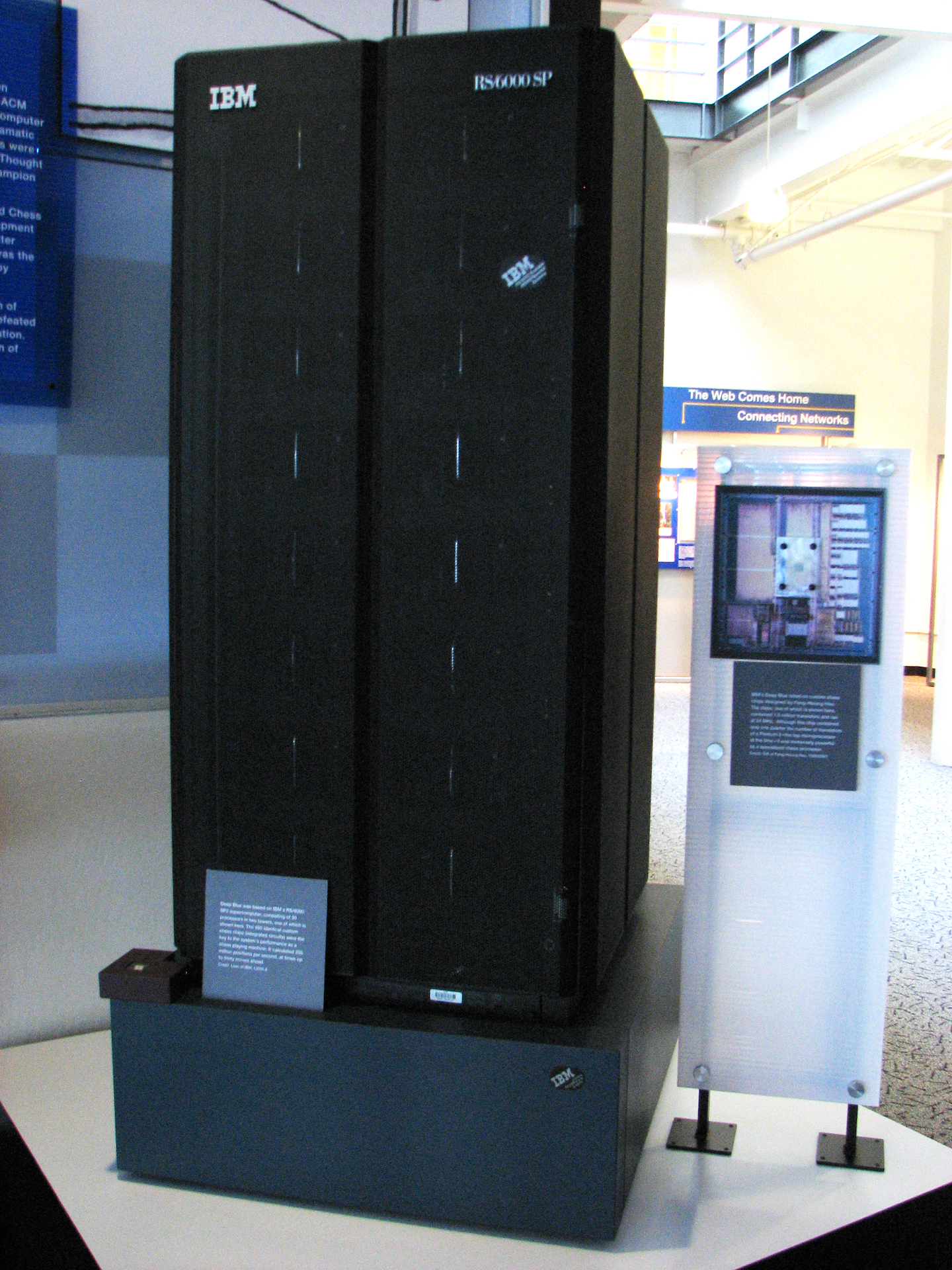 a black, rectangular object with a white "IBM" label in the corner sits atop a stand beside a plaque describing it