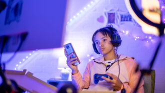 A photo of a teen sitting in front of their bedroom desk. They are looking at a monitor, listening to headphones, holding up a smartphone, and potentially streaming (there's a ring light that is turned on pointed at them).