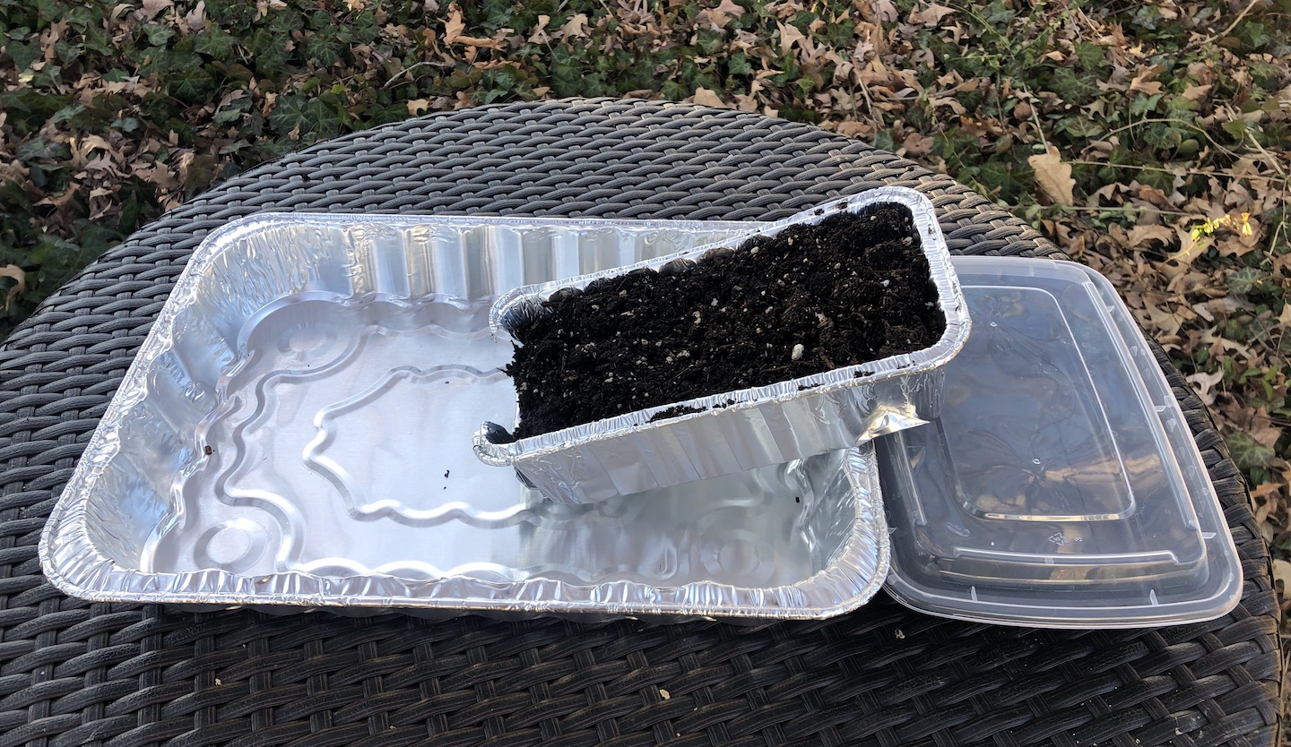 one end of a bread pan filled with soil sits in a cake pan, while the other end is propped up on a plastic container