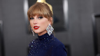 Taylor Swift is looking at the viewer.Her hair is up and she is wearing huge diamond and purple gem earrings. with a navy blue outfit is covered in glittering beads.