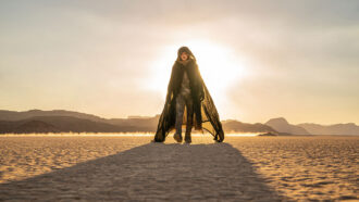 Actor Timothée Chalamet wears a brown cloak with a hood. He is walking toward the camera in a desert world with the sun shining behind him.