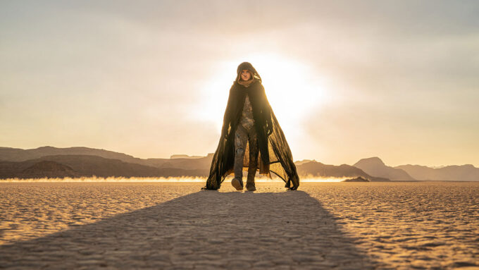 Actor Timothée Chalamet wears a brown cloak with a hood. He is walking toward the camera in a desert world with the sun shining behind him.