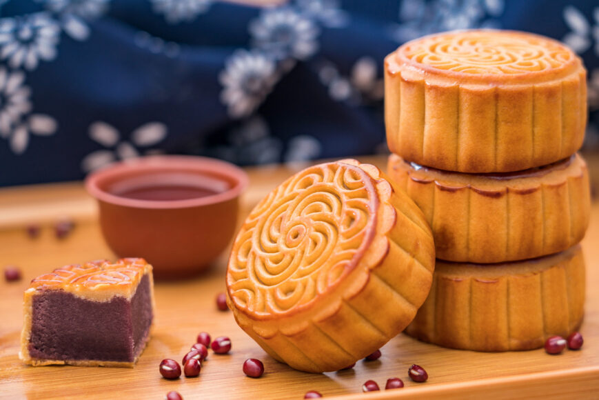 A stack of brown mooncakes sit on a wooden table. Red beans are scattered nearby.