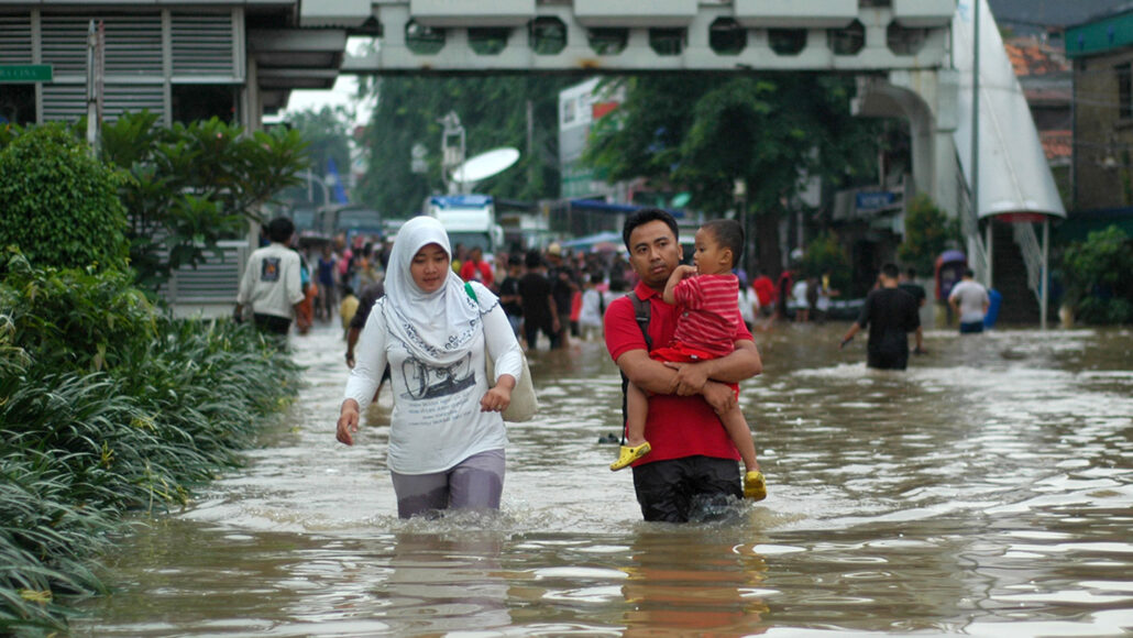 people walk through a flooded street in Indonesia, a woman and a man are walking toward the viewer in water that is thigh-high. The man is carrying a toddler.