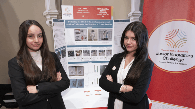 two teen girls stand side by side with their arms crossed in front of a science fair project poster