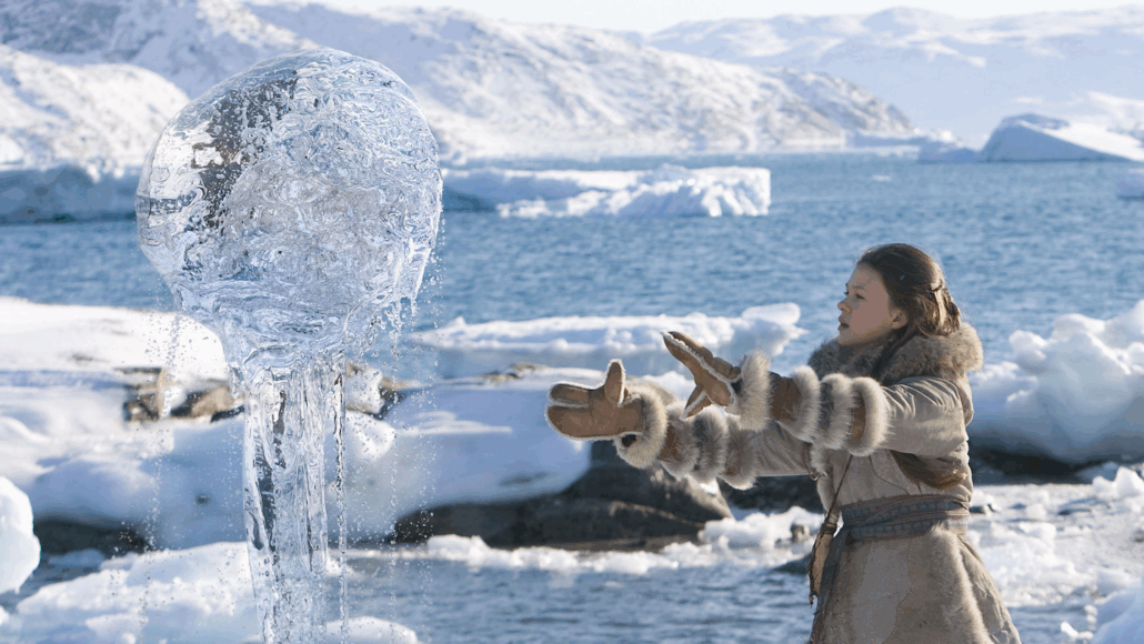 a girl in a parka, gloves and boots stands on a sheet of ice and snow with her hands out in front of her to levitate a ball of seawater into the air