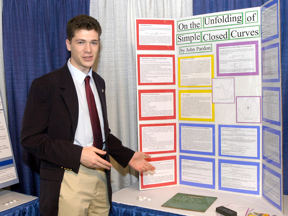 John Pardon, a teen wearing a suit, stands in front of a colorful posterboard showing his research