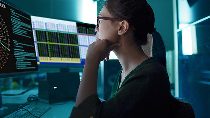a woman wearing glasses looking at multiple computer monitors in the dark
