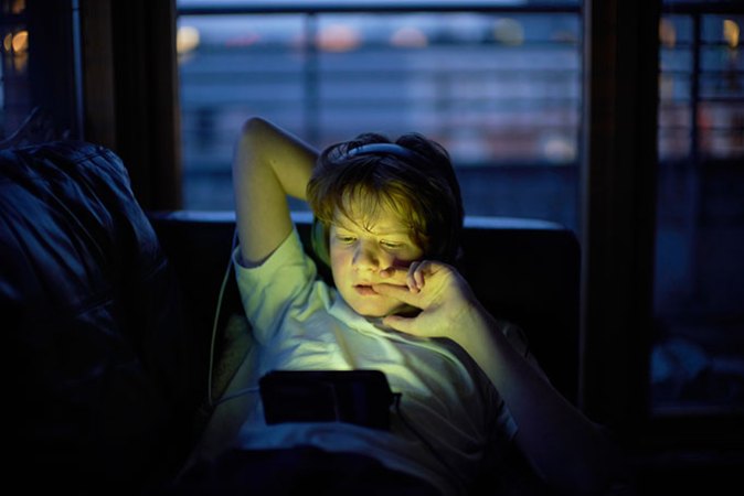 a kid lounging on a sofa and watching something on his tablet while wearing headphones
