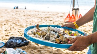 a photo of a beach. in the foreground are hands holding a blue mesh strainer full of plastic debris from the ocean