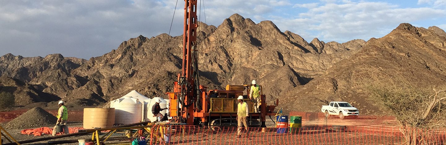 a large orange drill is in center of the picture. there are workers around the drilling site and mountains in the distance at the horizon.