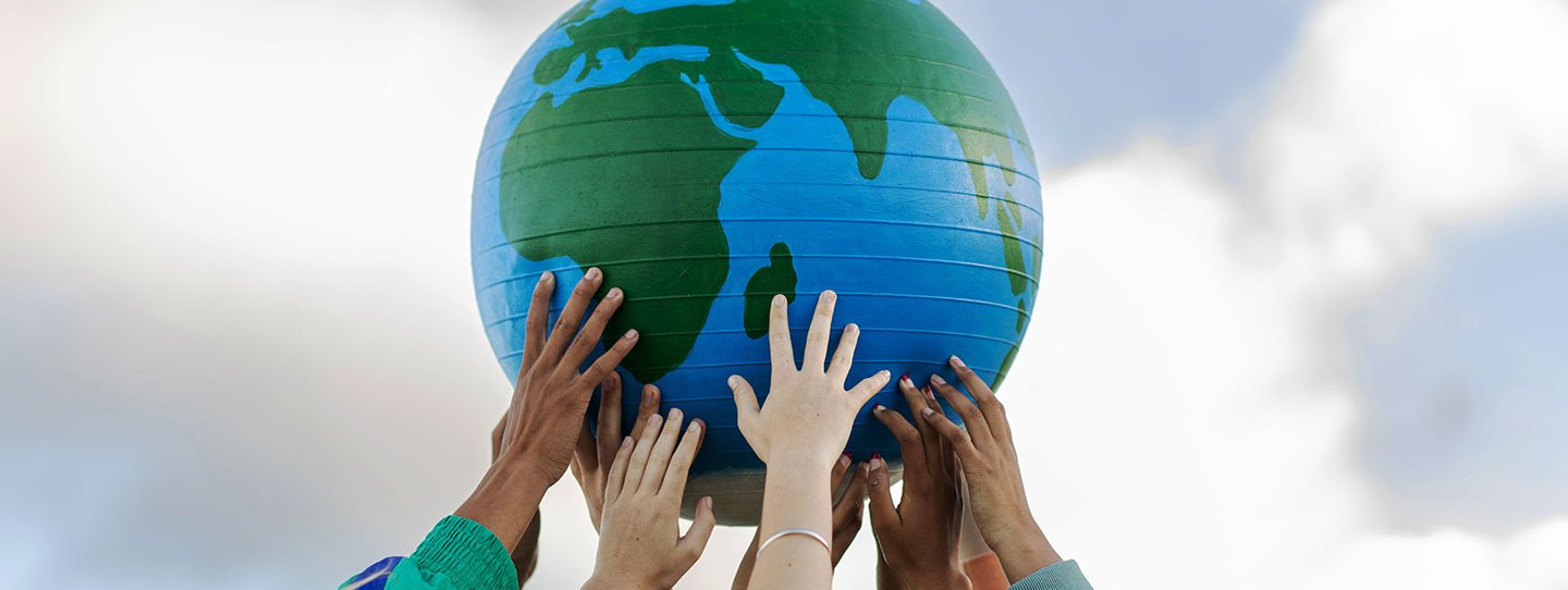 a photo of a giant ball painted to resemble the Earth. A group of hands of varying skin tones is holding the ball up to a cloudy gray sky.