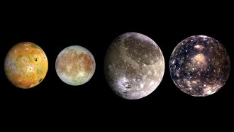 four pockmarked orbs of different sizes are in a row against a black background; the leftmost one (Io) is a splotchy orange-ish green; the second from right (Europa) is bluish-white with deep red scratches and patches; the second from right (Ganymede) is larger than the other two and silvery with white craters; the rightmost (Callisto) is slightly smaller than Ganymede and a deep blue with bright white spots