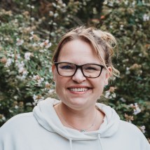 Megan Sever is standing in front of flowering bushes. She is blond with glasses.