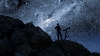 A person with a telescope standing on a dark hillside, silhouetted against a starry night sky.