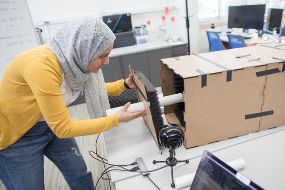 a photo of Shimaa Ahmed, a light brown skinned woman wearing a pattened headscarf, inserting the tube into a cardboard tunnel with sound absorbing foam lining the inside