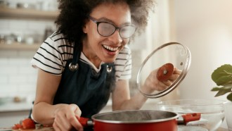 A young woman with curly hair and steamed glasses is opening the lid of a red pot.