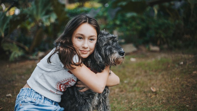 A brunette teen girl in a gray shirt and jeans kneels on the ground to hug a fluffy grey dog