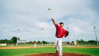 A baseball pitcher wearing a red jersey and white pants pitches a ball from a brown mound. A green baseball field surrounds him below cloudy skies
