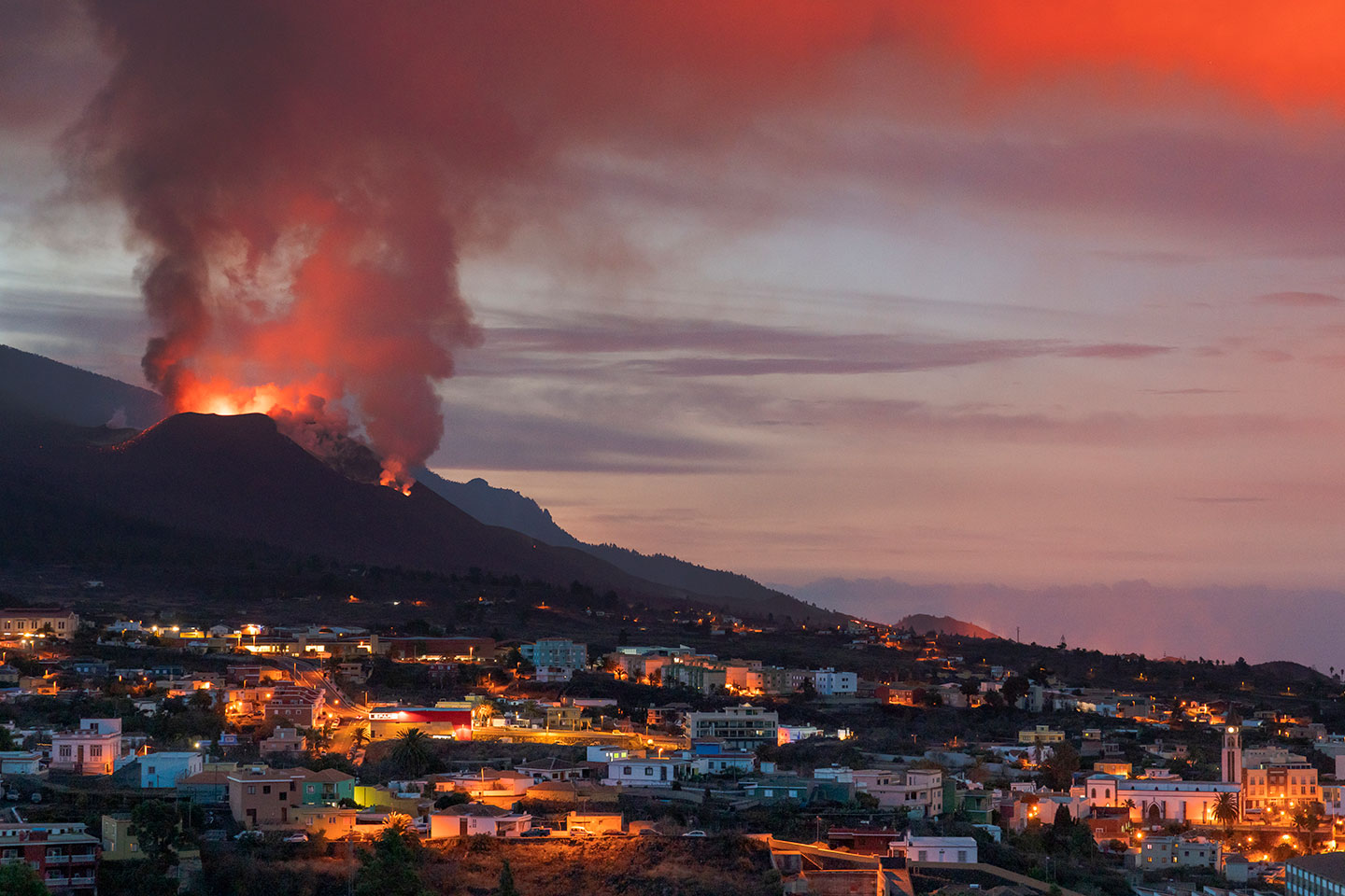 a photo from a distance, showing a volcano shooting ash and lava up into the air. In front of the volcano is city.