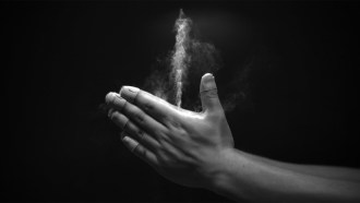 A grayscale images shows two hands clapping together, with a white jet of baby powder streaming upward from a space between the thumbs and forefingers.