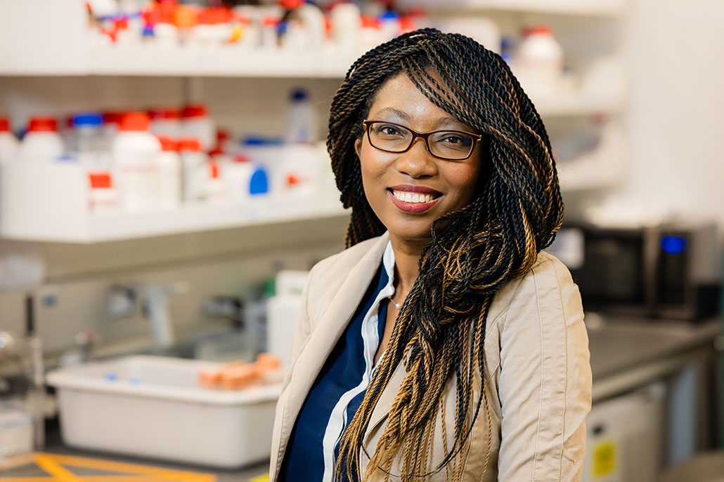 a photo of Ludmilla Aristilde,  a smiling adult woman with medium brown skin wearing a lab coat and standing in her laboratory. Her hair is styled in long thin braids and she wears glasses..