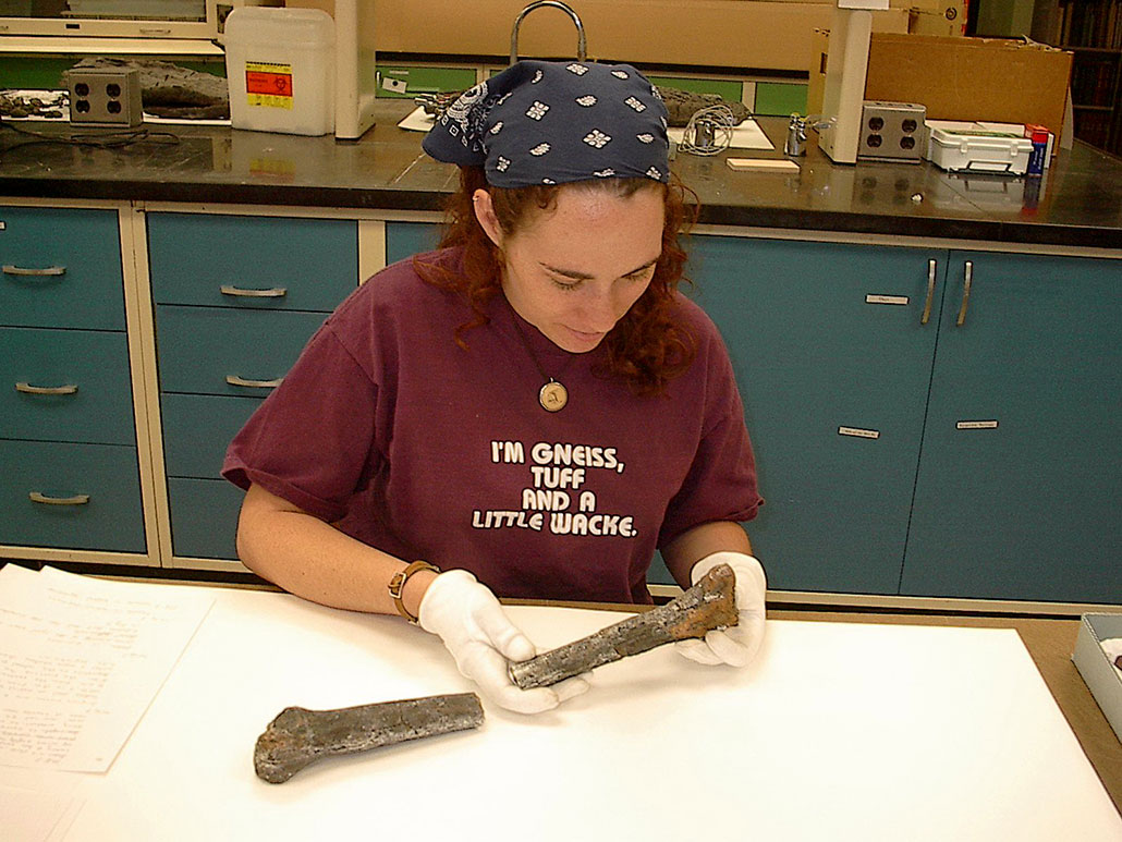 Paleontologist ReBecca Hunt-Foster is seated at a white table. She's holding a long brown fossil. She is wearing a navy bandana.