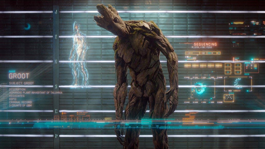 a still from the Guardians of the Galaxy movie, showing the treelike alien Groot standing with his head cocked to the left, examining a futuristic info screen