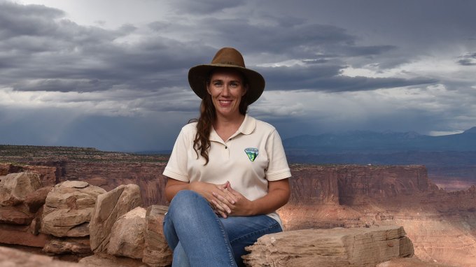 Paleontologist ReBecca Hunt-Foster sits on a stone formation. Dark grey clouds hang over a canyon behind her.