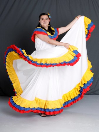 A. Catalina Vélez-Ortega wears a Cumbia outfit. The dress si white with yellow, blue and red frills. She has black hair and fair skin.