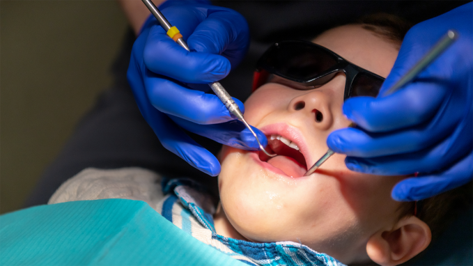 A child in sunglasses has dental tools inserted in mouth by practitioner wearing blue gloves.