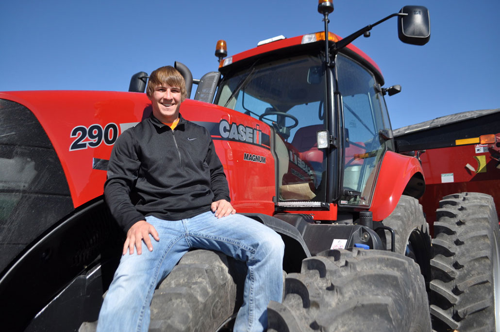 a photo of a young Mitchell Hora, a smiling kid with pale skin and wheat colored hair sitting on a tractor wheel and beaming