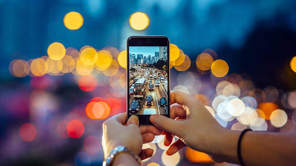 a photo of a cell phone taking a photo of a highway overpass (looking down at traffic) at dusk. The image on the phone is in focus, but the surroundings are out of focus.