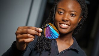 a photo of Paula Kirya holding a blue morpho butterfly wing and smiling. She has dark hair and dark skin.