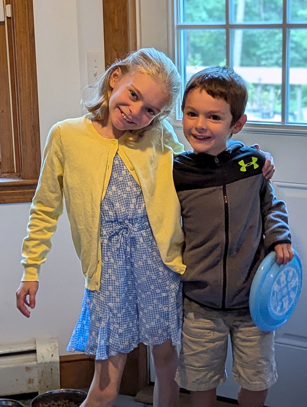 a 10 year old girl cocks her head to the side and smiles while hugging her little brother. She has pale skin, and short light blond hair, and is wearing a blue and white floral patterned dress with a yellow cardigan