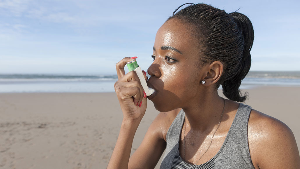 a woman is using an inhaler at a beach, she has dark skin and dark hair in microbraids and a ponytail. She seems to have been excercising.